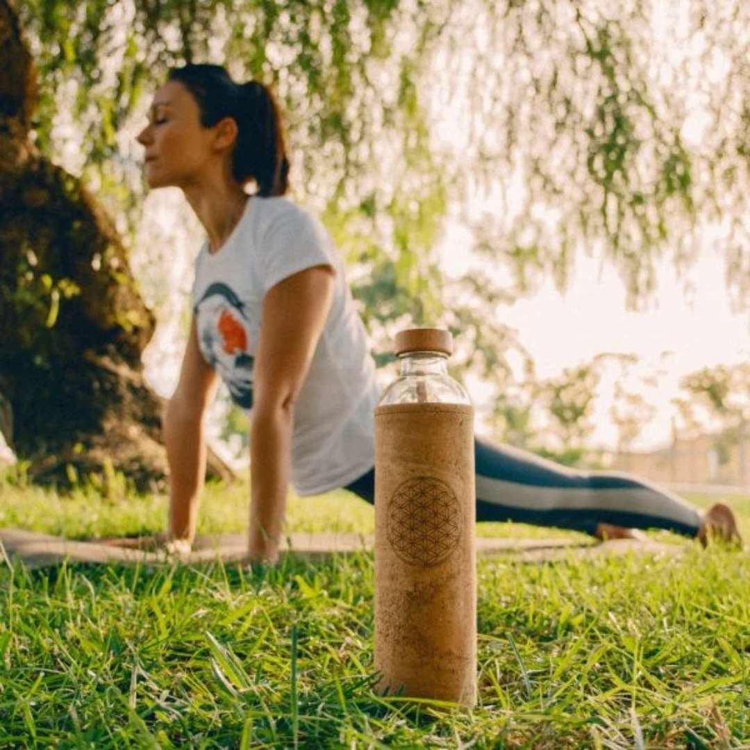 Flaska glass water bottle with cork sleeve (Flower of Life) on grass; woman practicing outdoor yoga in the background